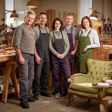 Fünfköpfiges Handwerksteam in der Werkstatt zwischen Holzböcken und einem restaurierten Sessel